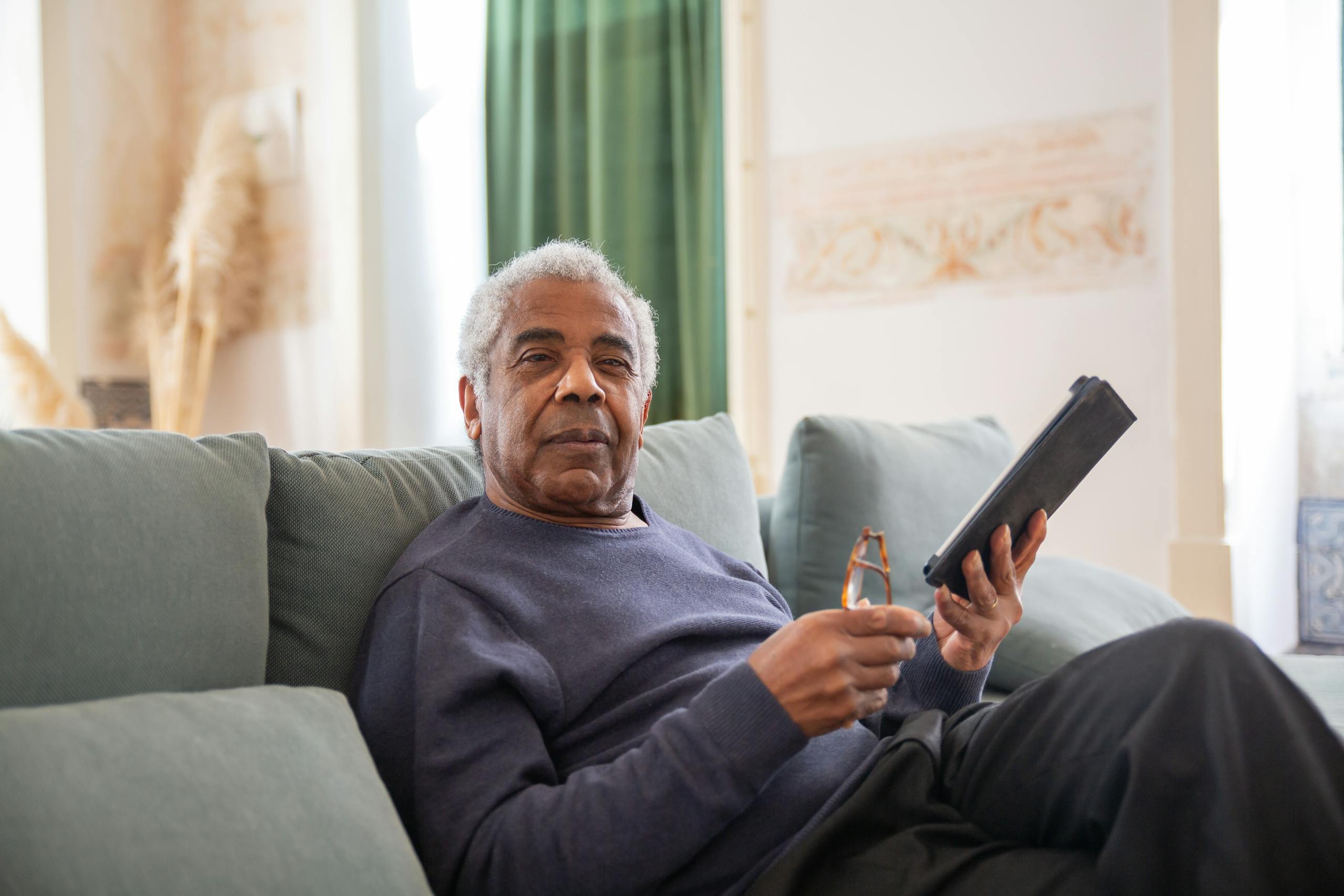 Senior African American man sitting on couch with digital tablet and eyeglasses, looking at camera. Retired Professional